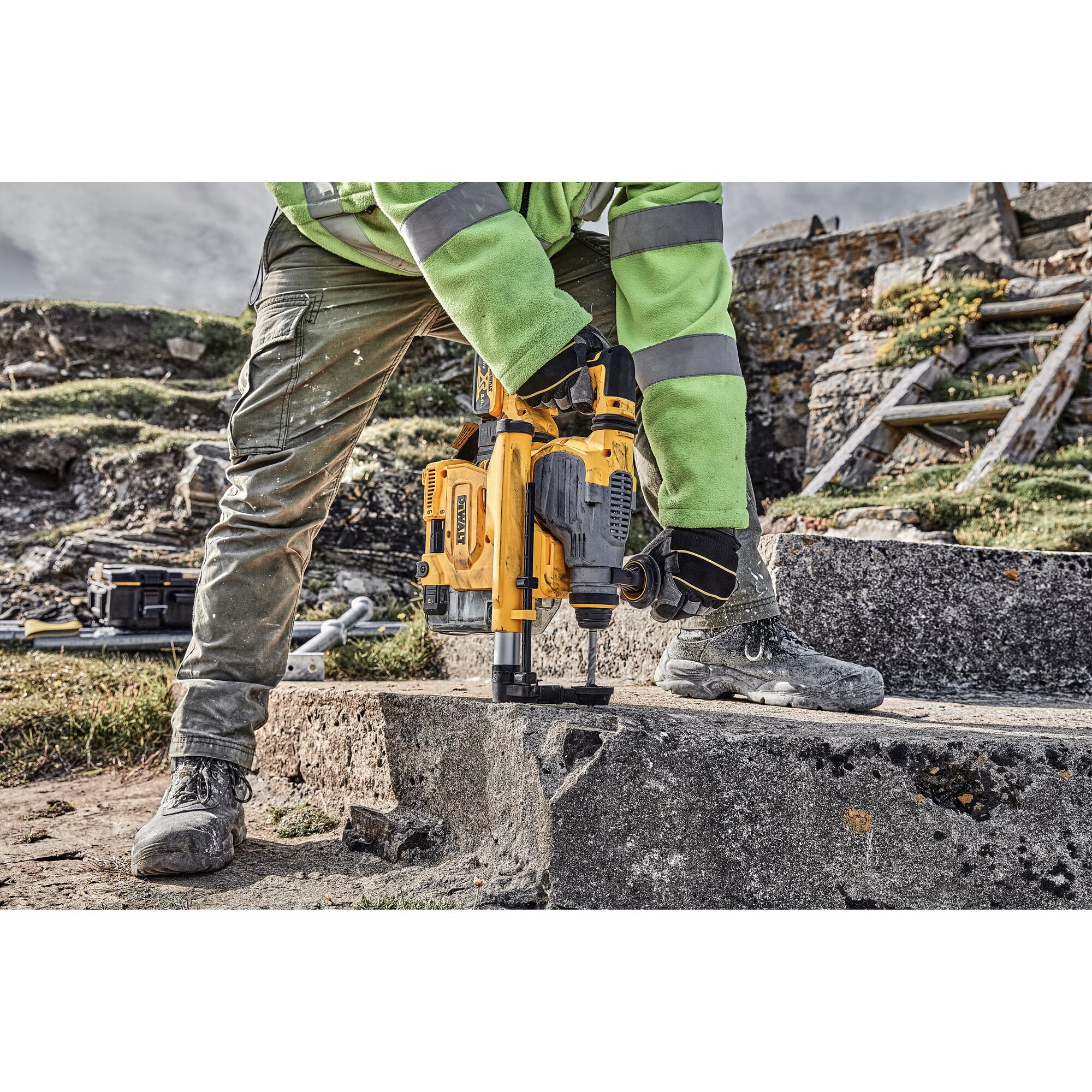 A person wearing work clothes and safety gloves is using a yellow DEWALT rotary hammer drill on a concrete surface outdoors. The drill is equipped with a dust collection attachment.