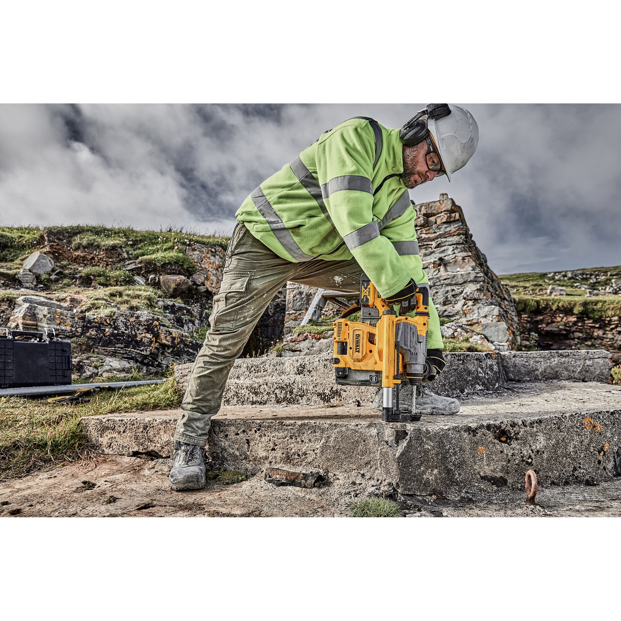 A construction worker wearing a green reflective jacket, protective helmet, and ear protection is using a yellow DEWALT drill on a concrete surface outdoors.