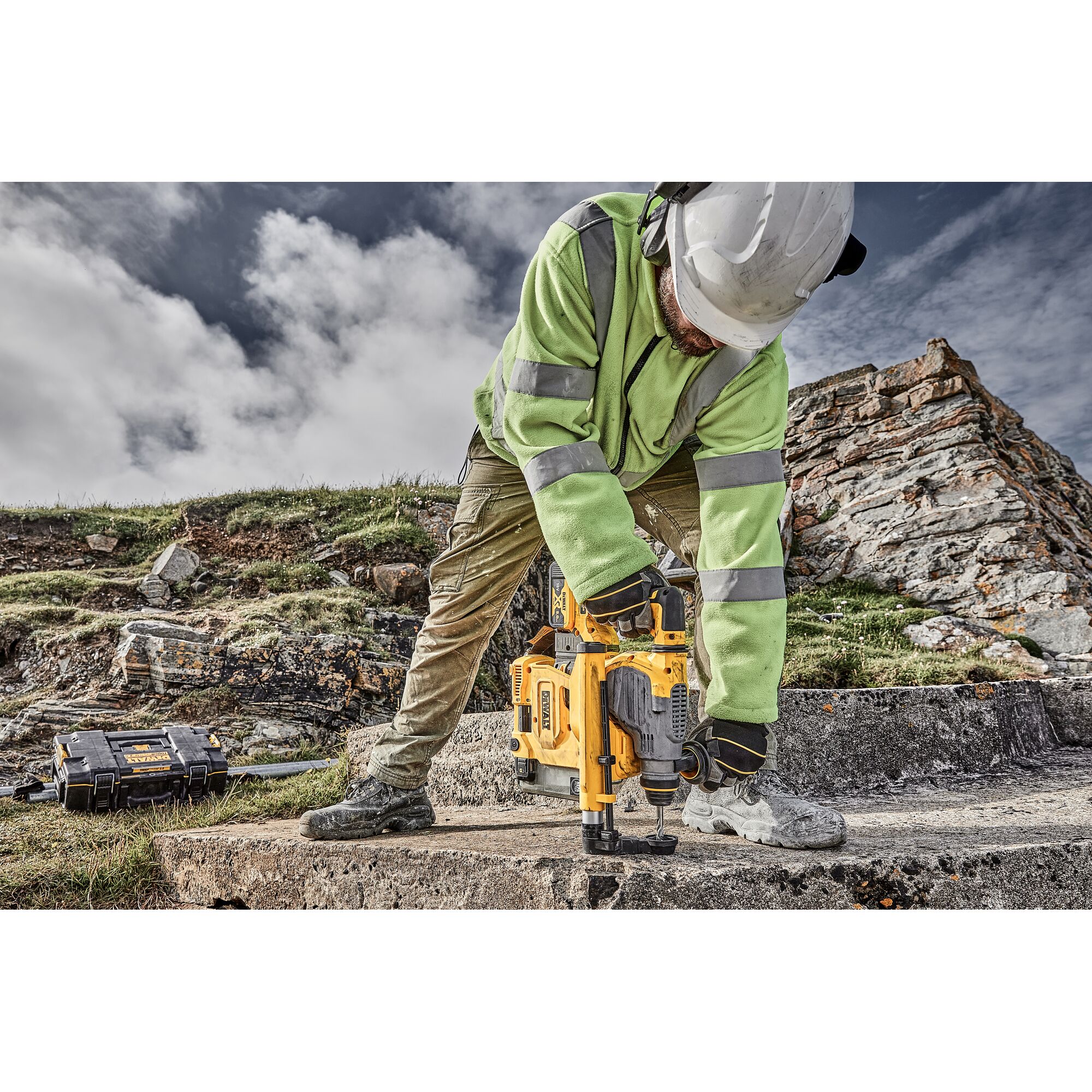 A worker wearing a hard hat and high-visibility jacket operates a yellow DEWALT rotary hammer drill on a concrete surface outdoors, with rocky terrain and a tool case visible in the background.