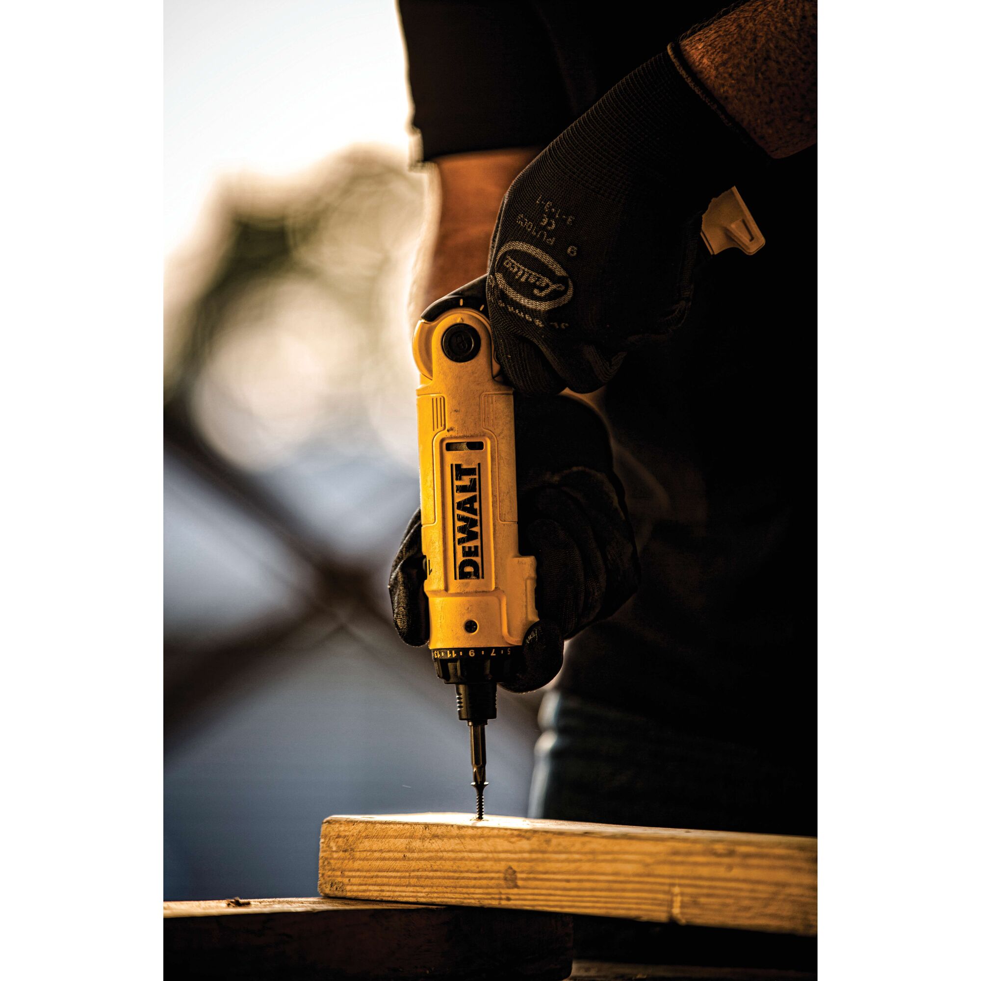 Close-up of a person wearing black gloves using a yellow DEWALT cordless screwdriver to drive a screw into a piece of wood.