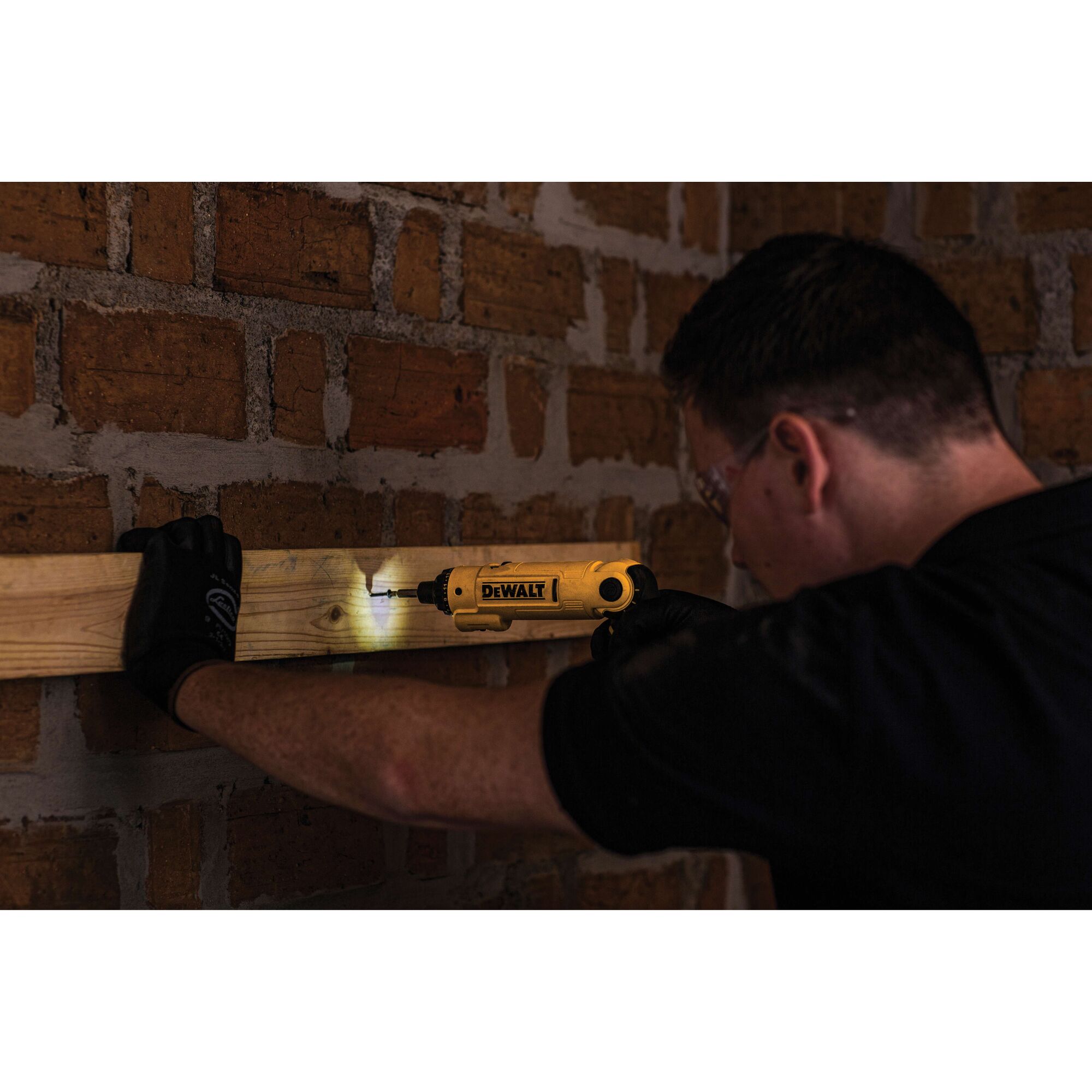 A person using a DEWALT DCF008 power tool to drive a screw into a wooden beam attached to a brick wall. The tool has an LED light to illuminate the work area. The person's face is blurred and they are wearing a black shirt and gloves.