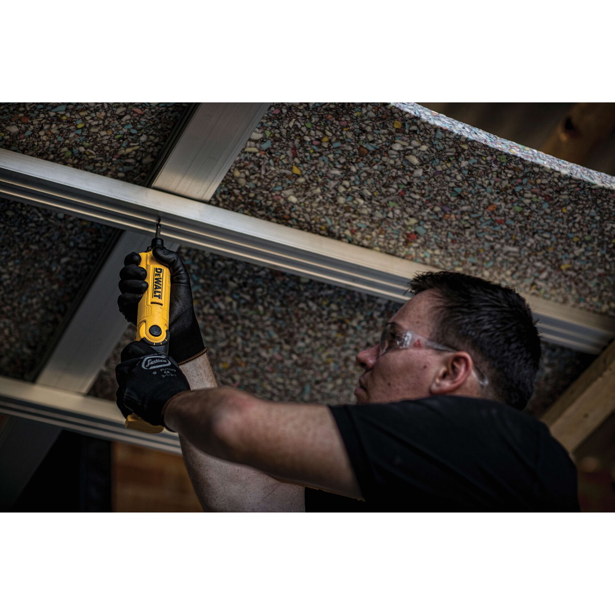 A person wearing gloves is using a yellow DEWALT DCF008 power tool to fasten a screw into a metal frame overhead. The person's face is blurred for privacy.
