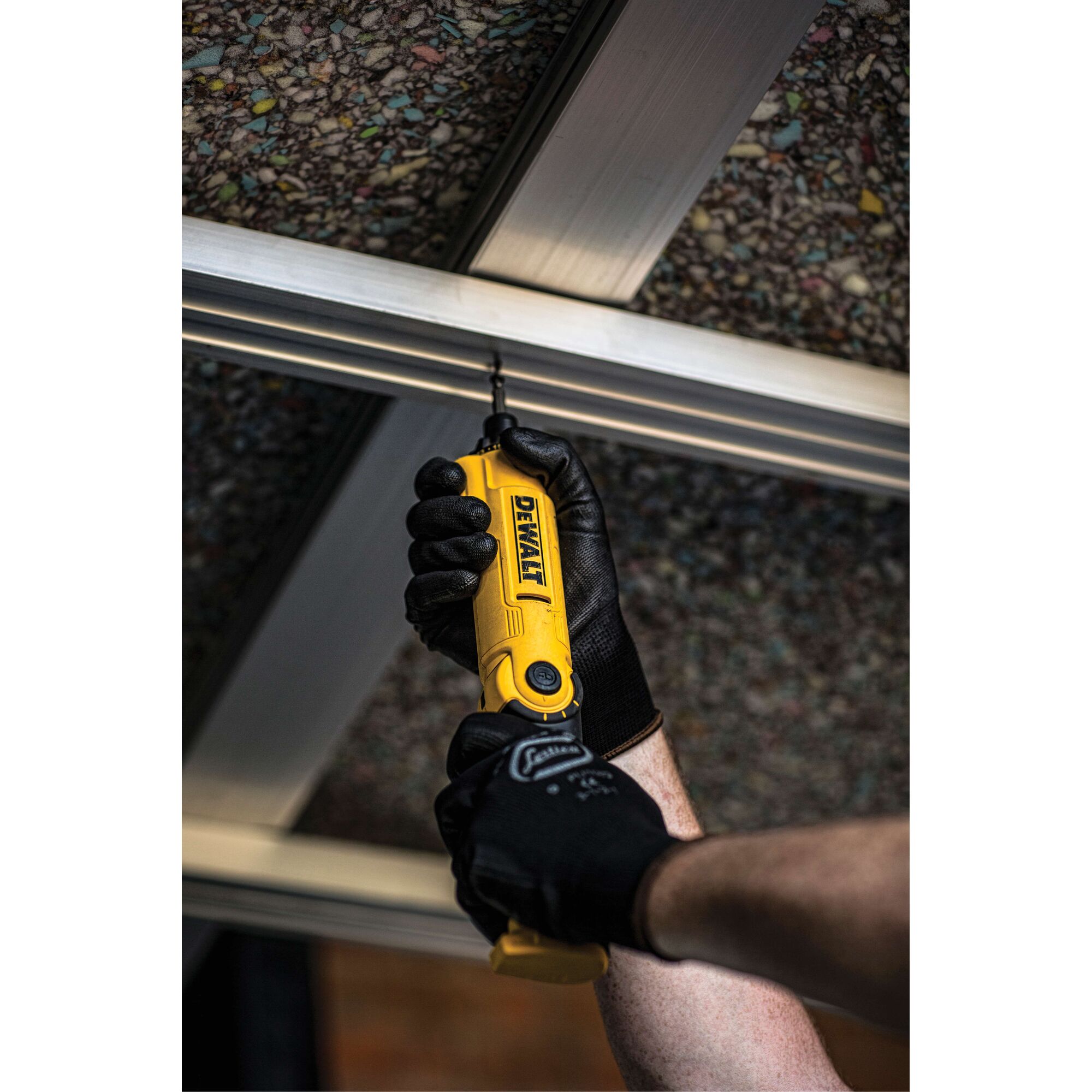 A person wearing black work gloves is using a yellow DEWALT cordless screwdriver to drill into a metal frame on the ceiling.