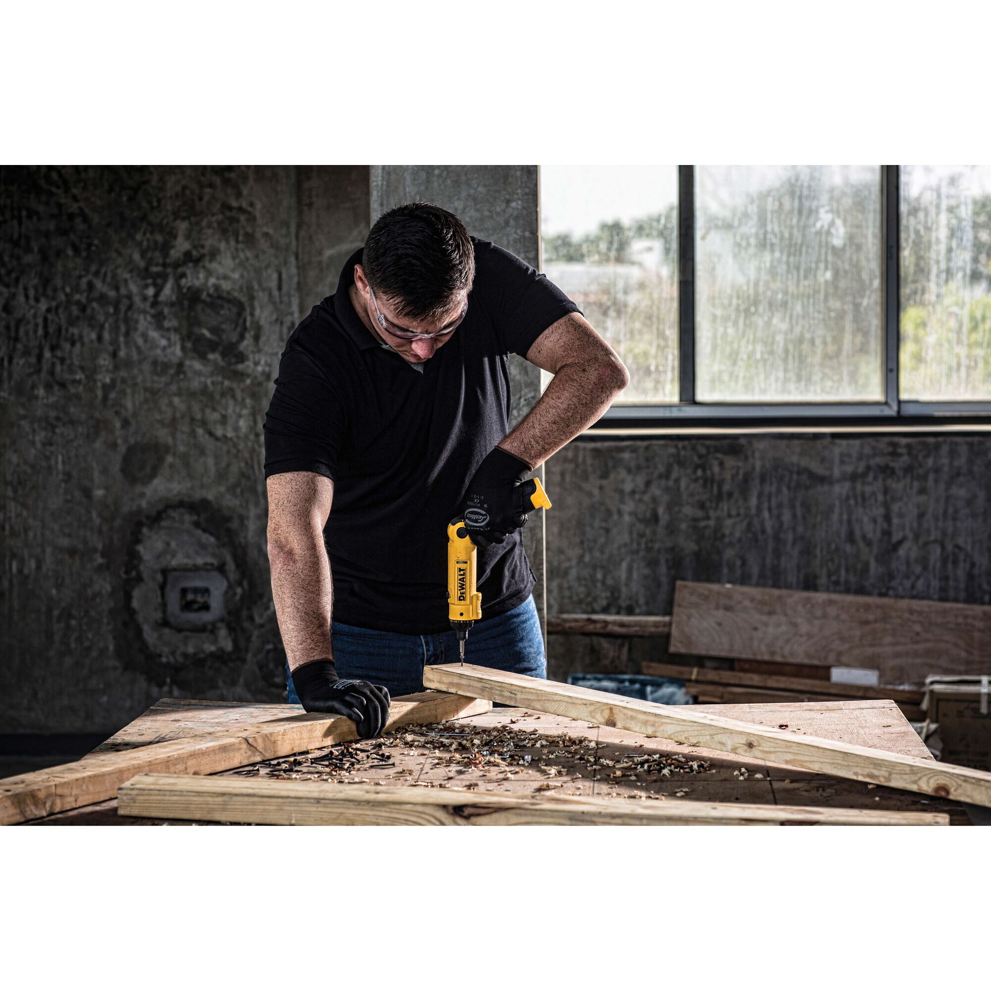 A person wearing black gloves uses a yellow DEWALT DCF008 cordless drill on a wooden frame in a workshop with concrete walls and a window.