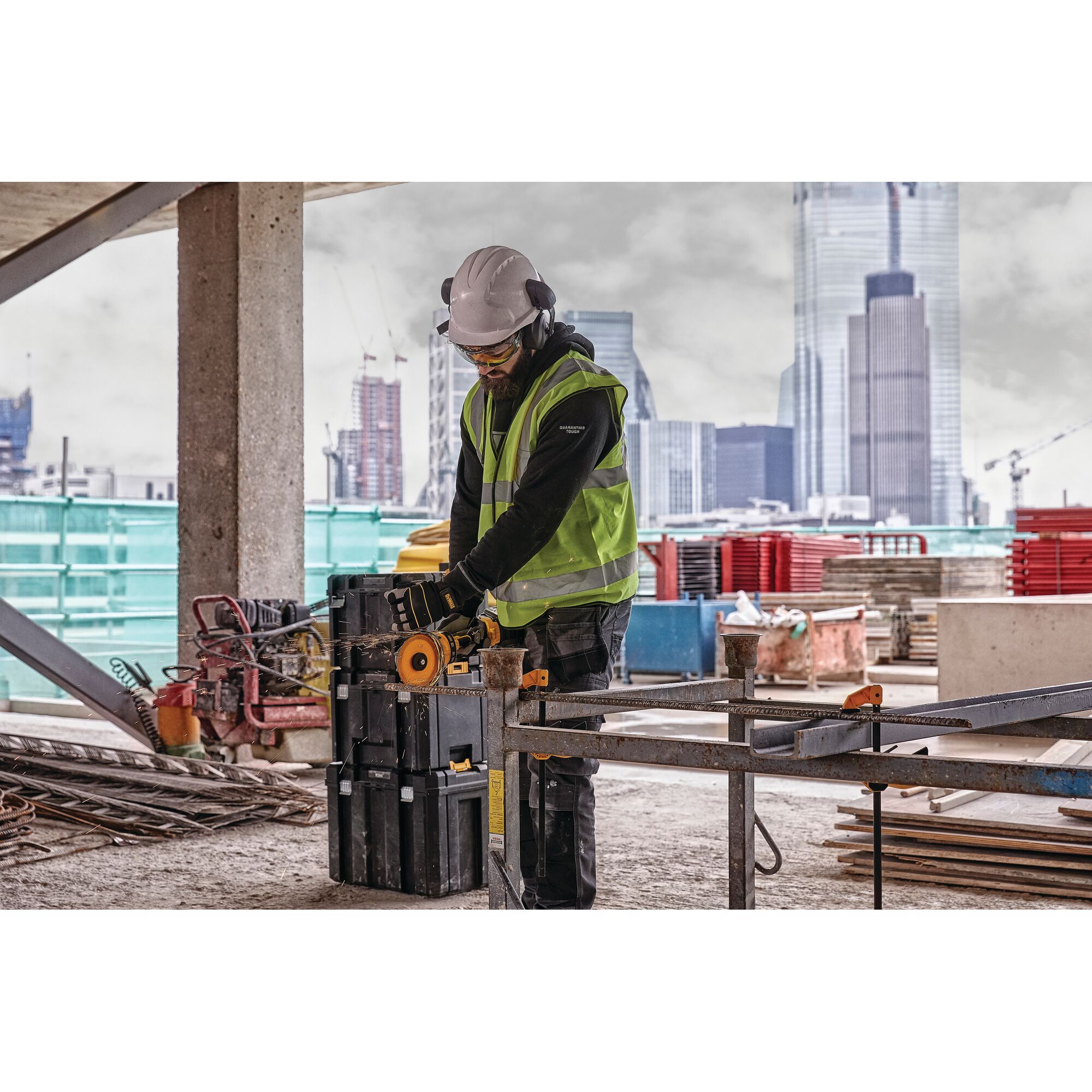 A construction worker wearing a high-visibility vest, helmet, ear protection, and gloves is using a DEWALT power tool to cut metal on a construction site. Tall buildings and construction materials are visible in the background.