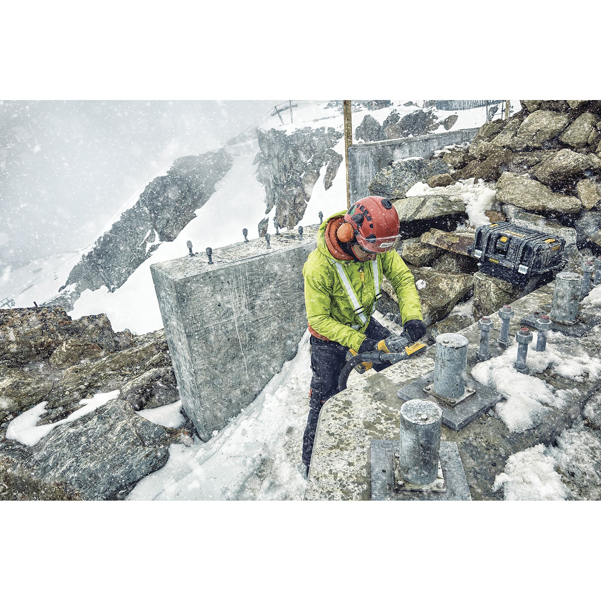 A worker wearing a helmet and winter gear uses a DEWALT power tool on a snow-covered mountain construction site with concrete and metal fixtures, surrounded by rocky terrain and snow.