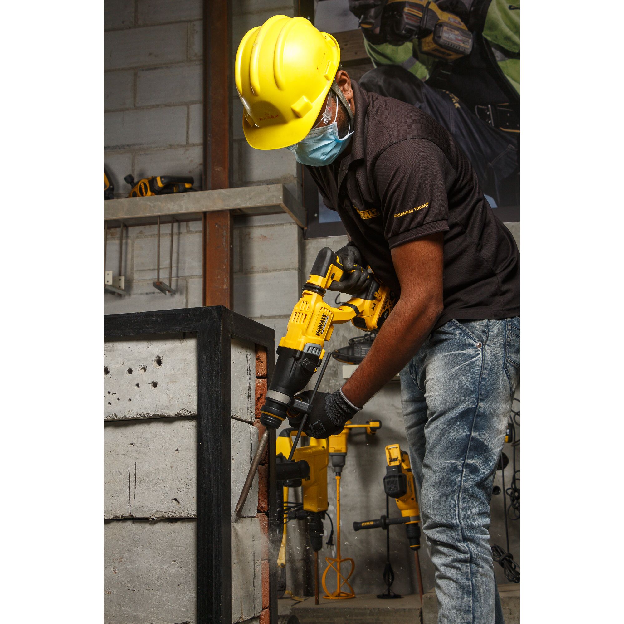 A worker wearing a yellow hard hat, protective glasses, and a face mask is using a DEWALT DCH263P2 power tool to drill into a concrete block in an industrial setting. Other DEWALT tools are visible in the background.