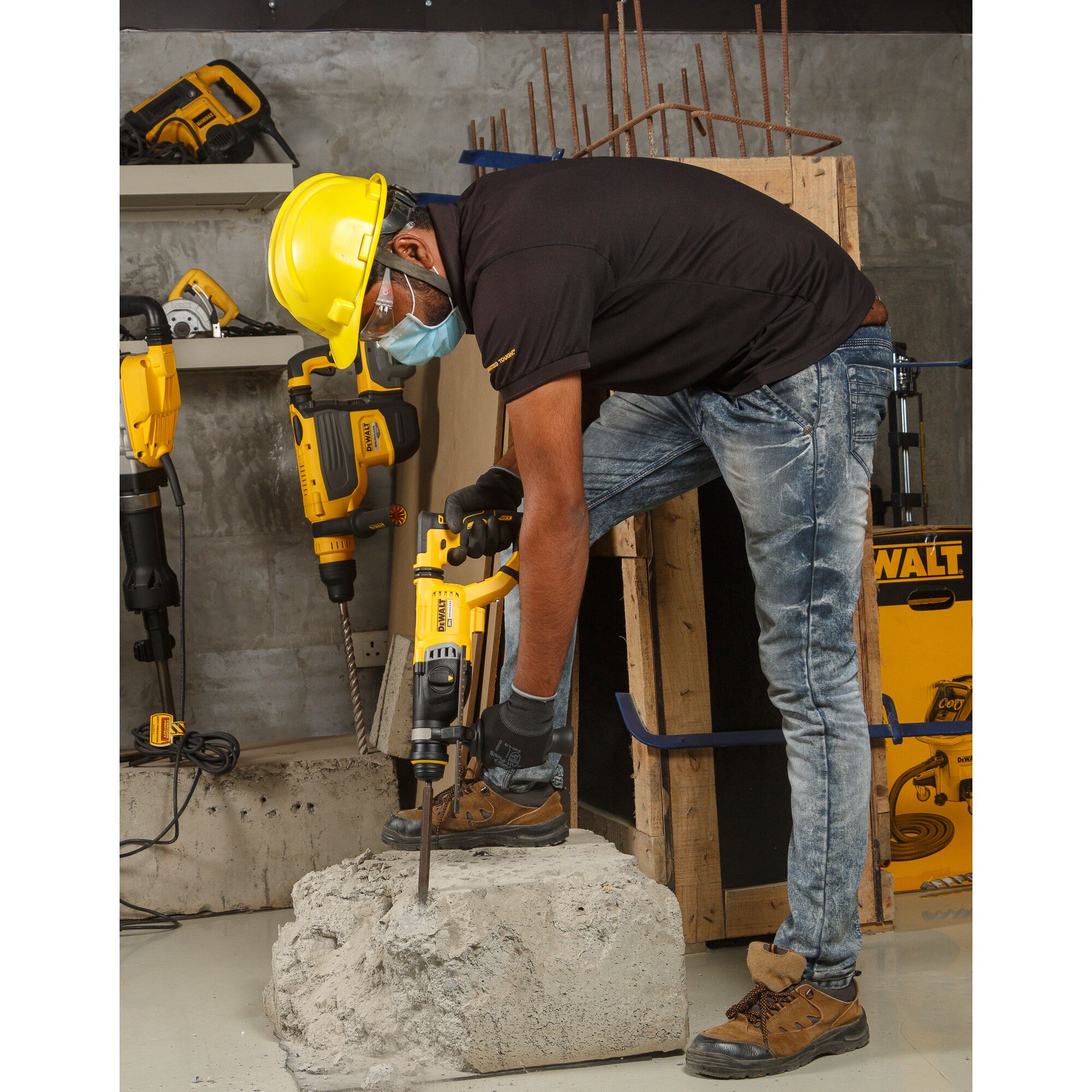 A person wearing a yellow safety helmet, face mask, gloves, and safety shoes is using a DEWALT rotary hammer drill (DCH263P2) to break a large concrete block in a construction setting. Various DEWALT tools and construction materials are visible in the background.