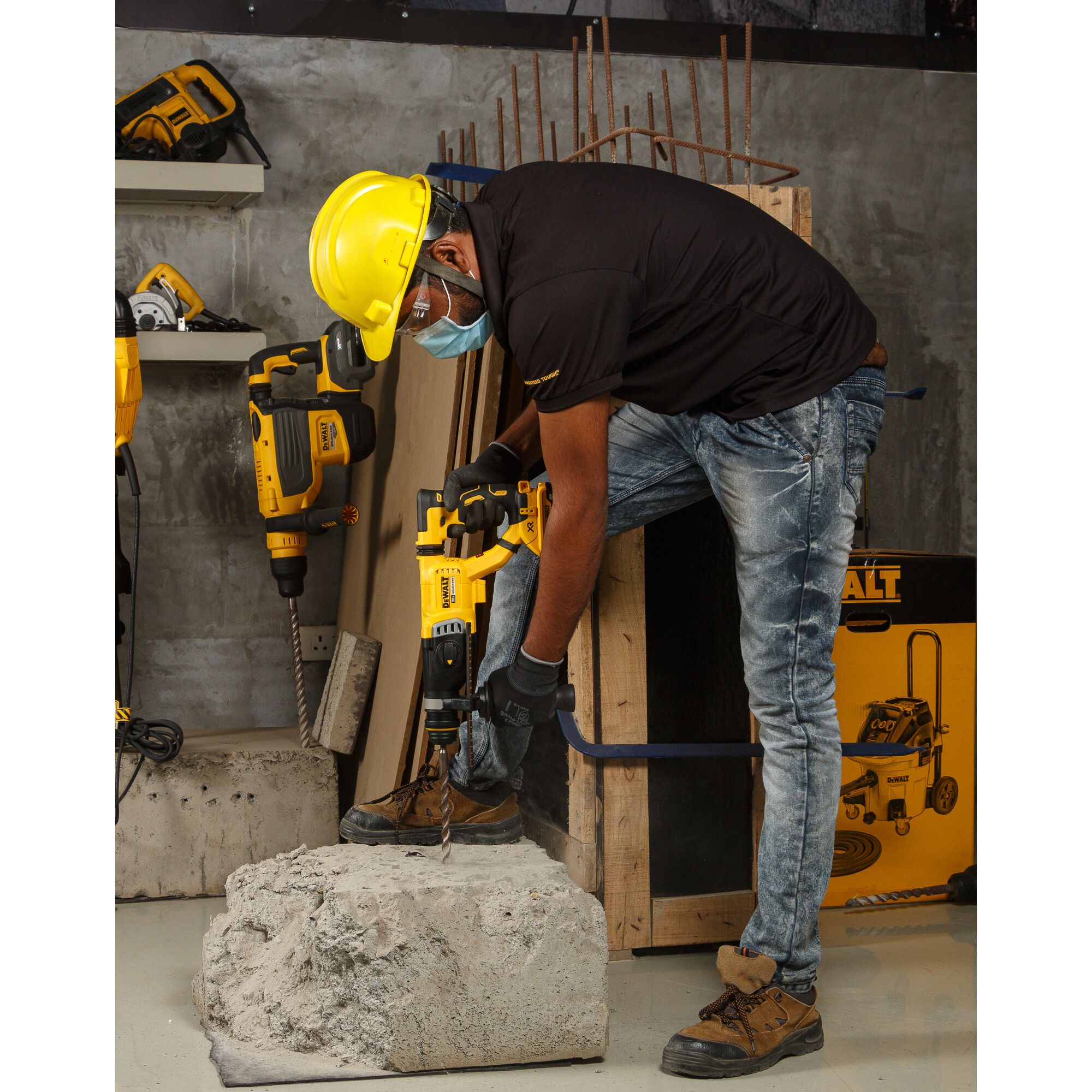 A person wearing a yellow safety helmet, face mask, gloves, and work boots is using a DEWALT rotary hammer drill to drill into a large piece of concrete in a workshop. The background shows more DEWALT tools and construction materials.