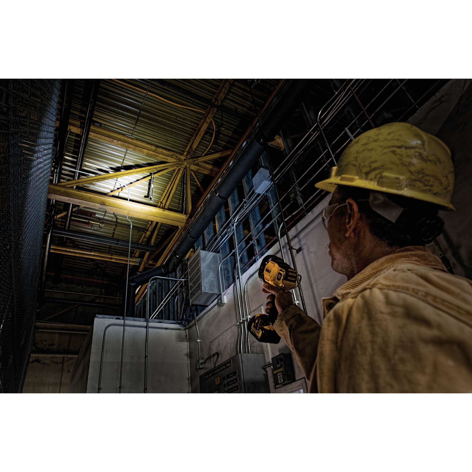 A worker wearing a yellow hard hat uses a DEWALT handheld spotlight to illuminate the ceiling and overhead pipes inside an industrial building.