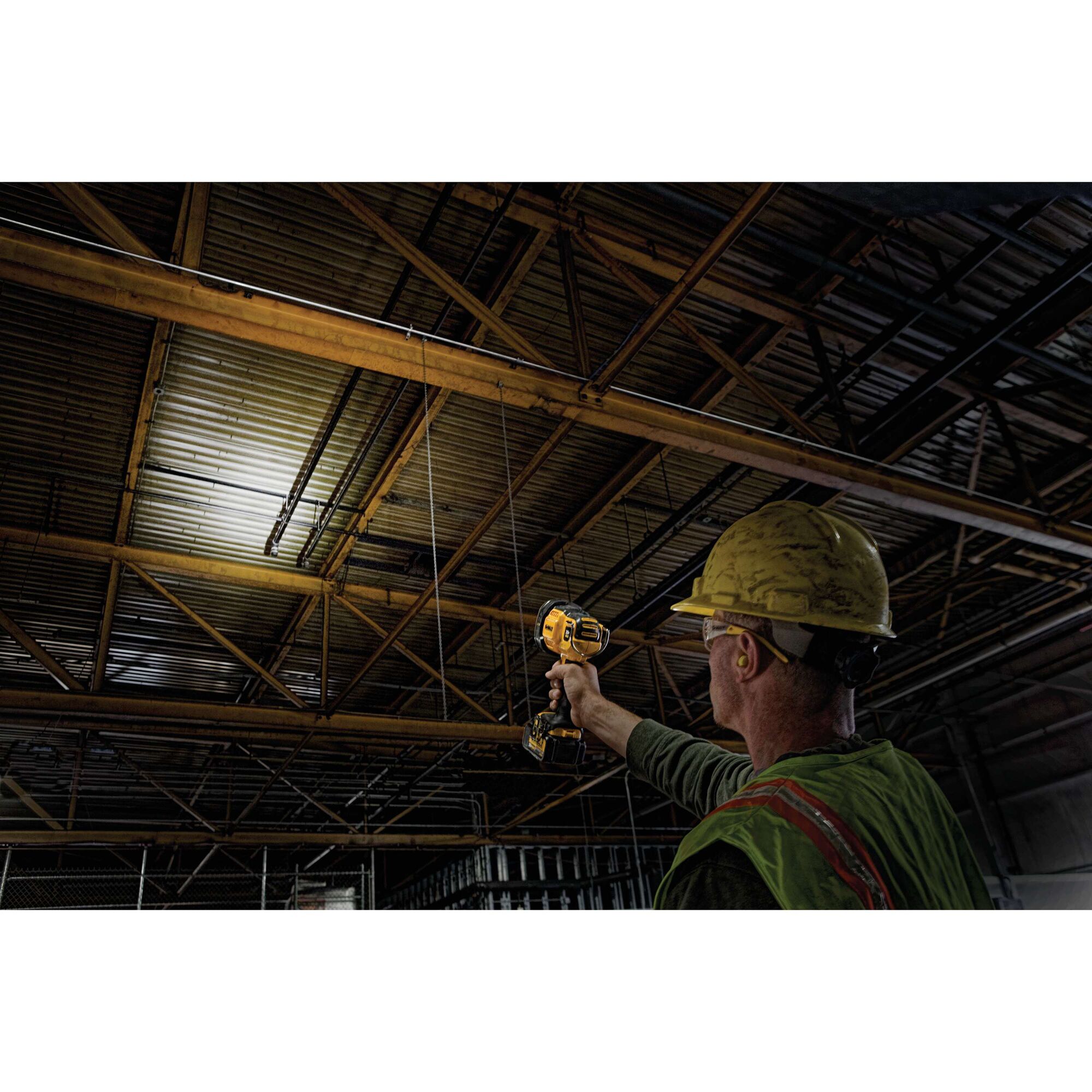 A person wearing a yellow hard hat and safety vest uses a DEWALT handheld spotlight to illuminate a metal ceiling inside an industrial building.