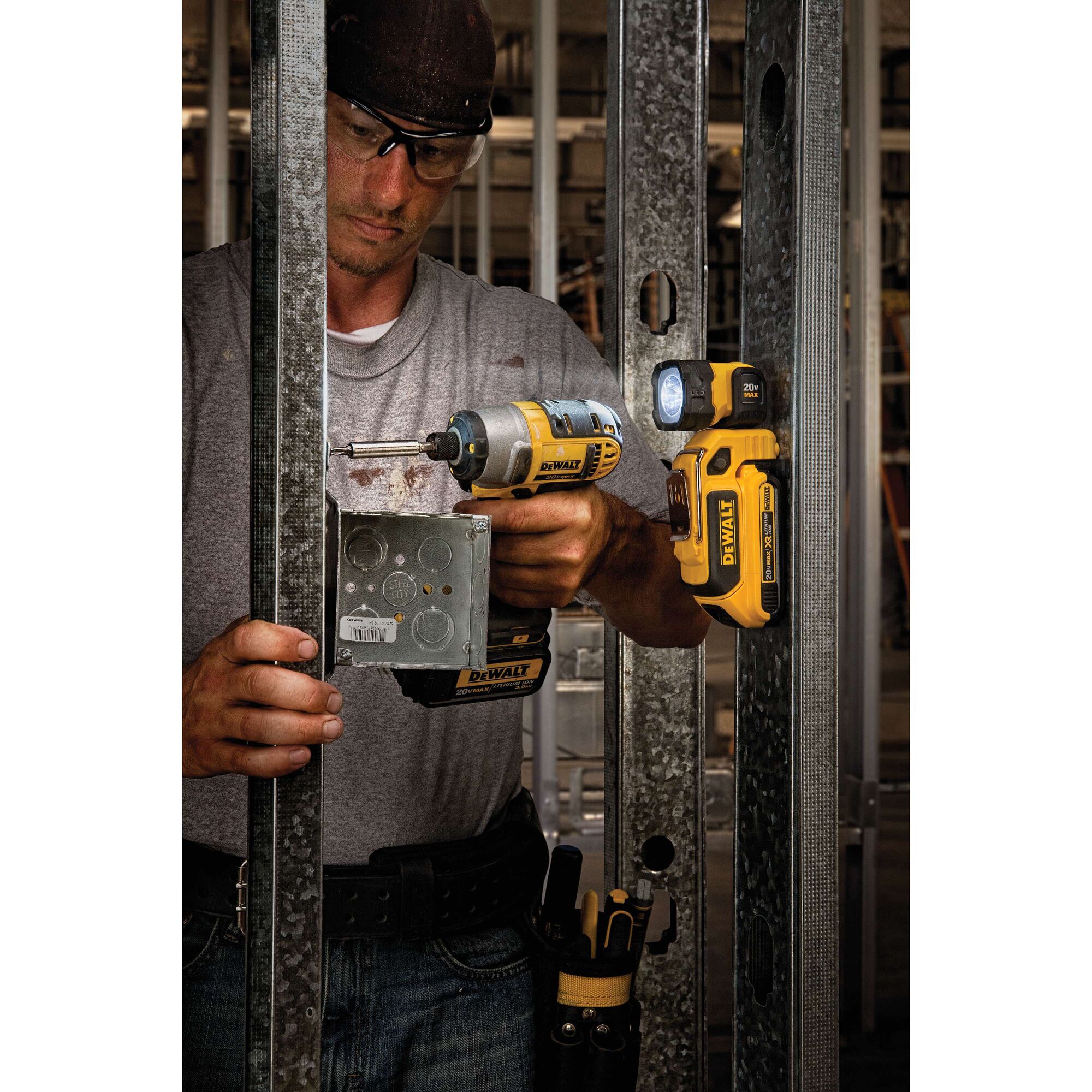 A person using a DEWALT power drill to install a metal electrical box into a steel framing structure. A DEWALT DCL044 portable LED work light is magnetically attached to the metal frame, illuminating the workspace. The person's face is obscured.