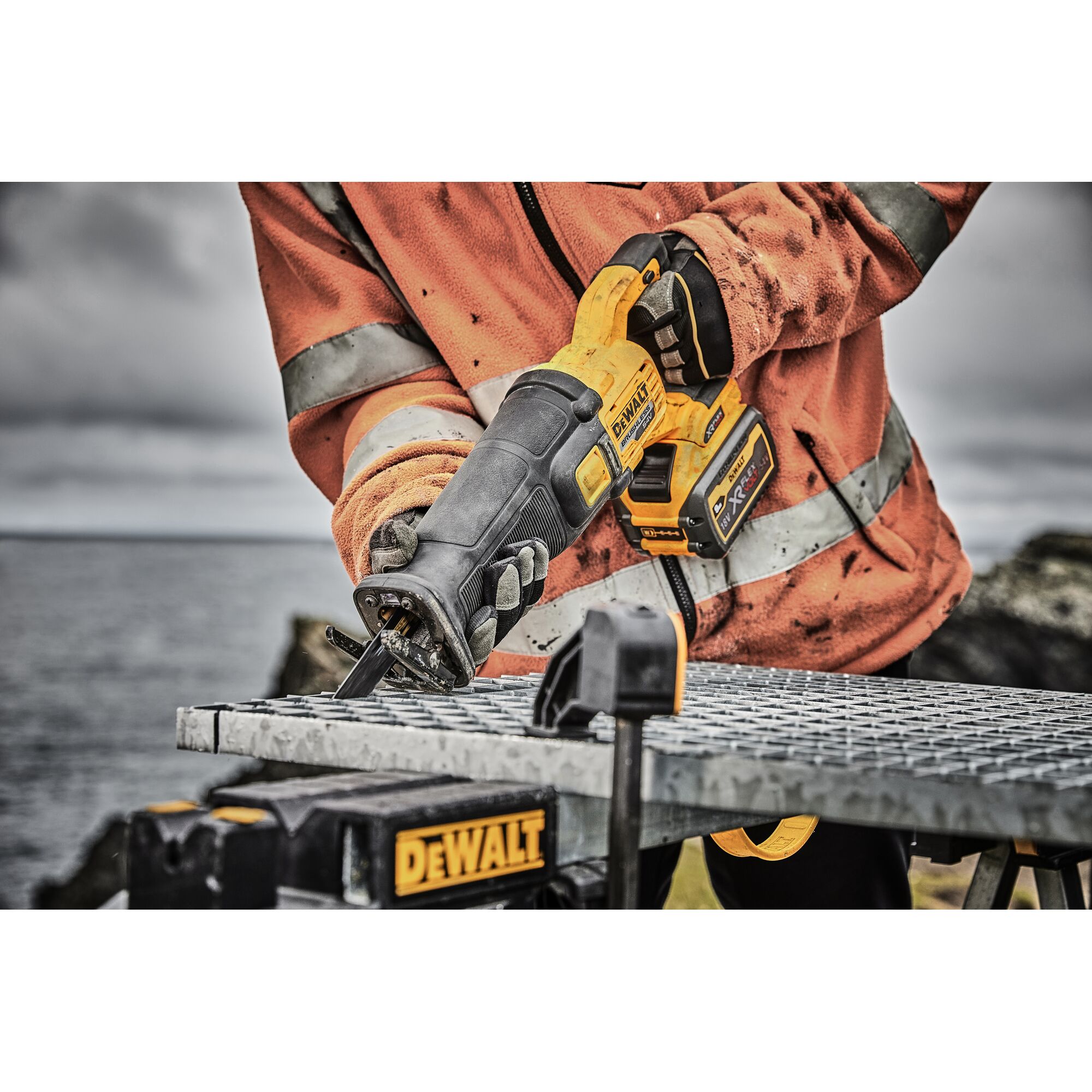 A person wearing an orange work jacket is using a DEWALT cordless reciprocating saw to cut through a metal grate outdoors. The DEWALT logo is visible on the saw and workbench.