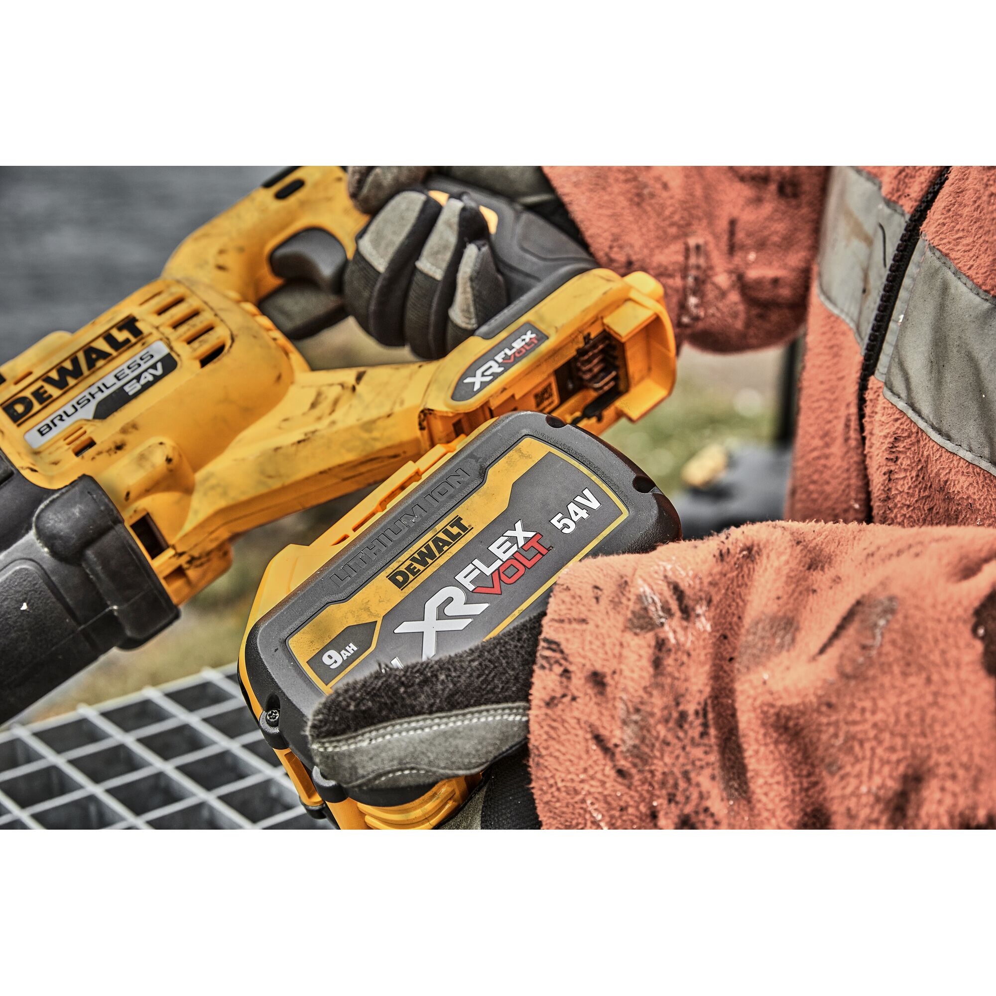 A close-up view of a worker holding a DEWALT XR FlexVolt 54V power tool, featuring a brushless motor and a removable lithium-ion battery. The worker is wearing gloves and a worn-out, orange safety jacket.