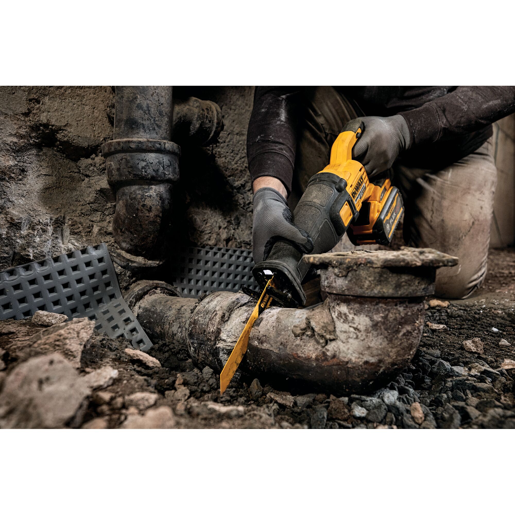 A person wearing work gloves uses a DEWALT cordless reciprocating saw to cut through an old metal pipe in a construction or plumbing setting. The scene shows rough terrain and industrial pipes.