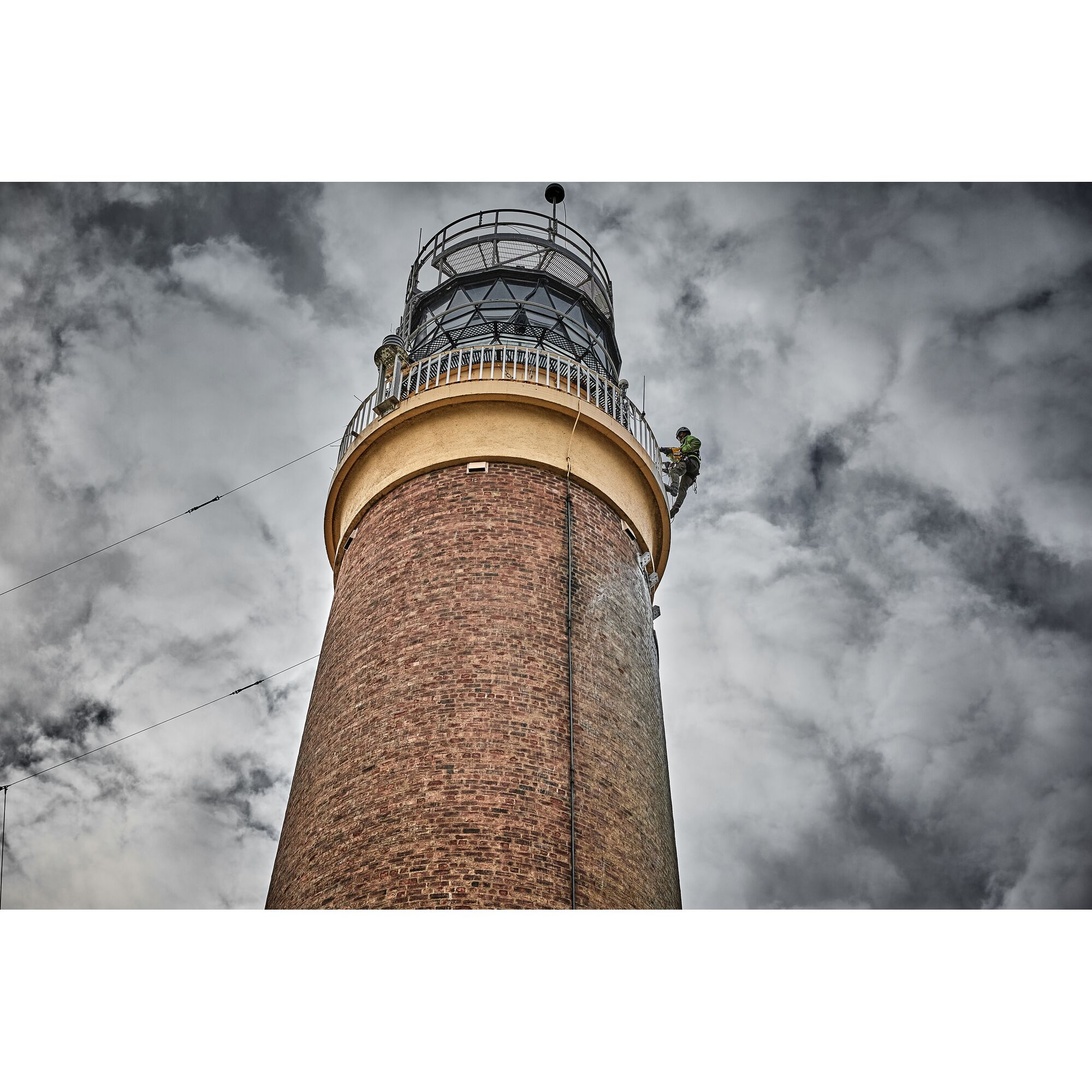 A tall brick lighthouse viewed from below against a cloudy sky, with a worker wearing safety gear ascending near the top of the structure.