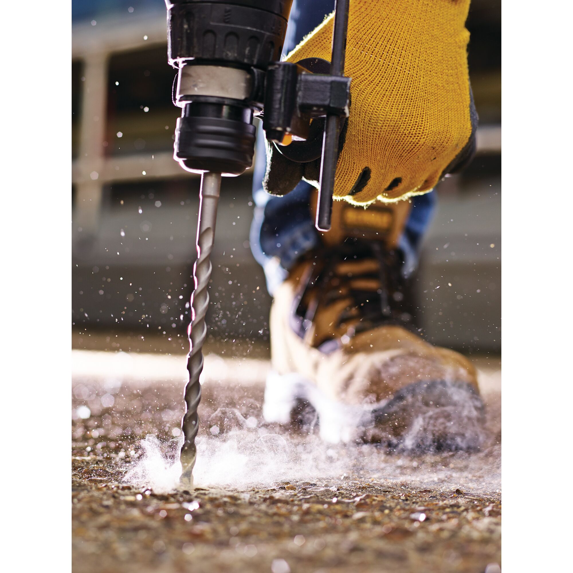 A close-up image of a person using a power drill with a masonry bit to drill into concrete, wearing yellow safety gloves and work boots. Dust and debris are visible around the drilling area.