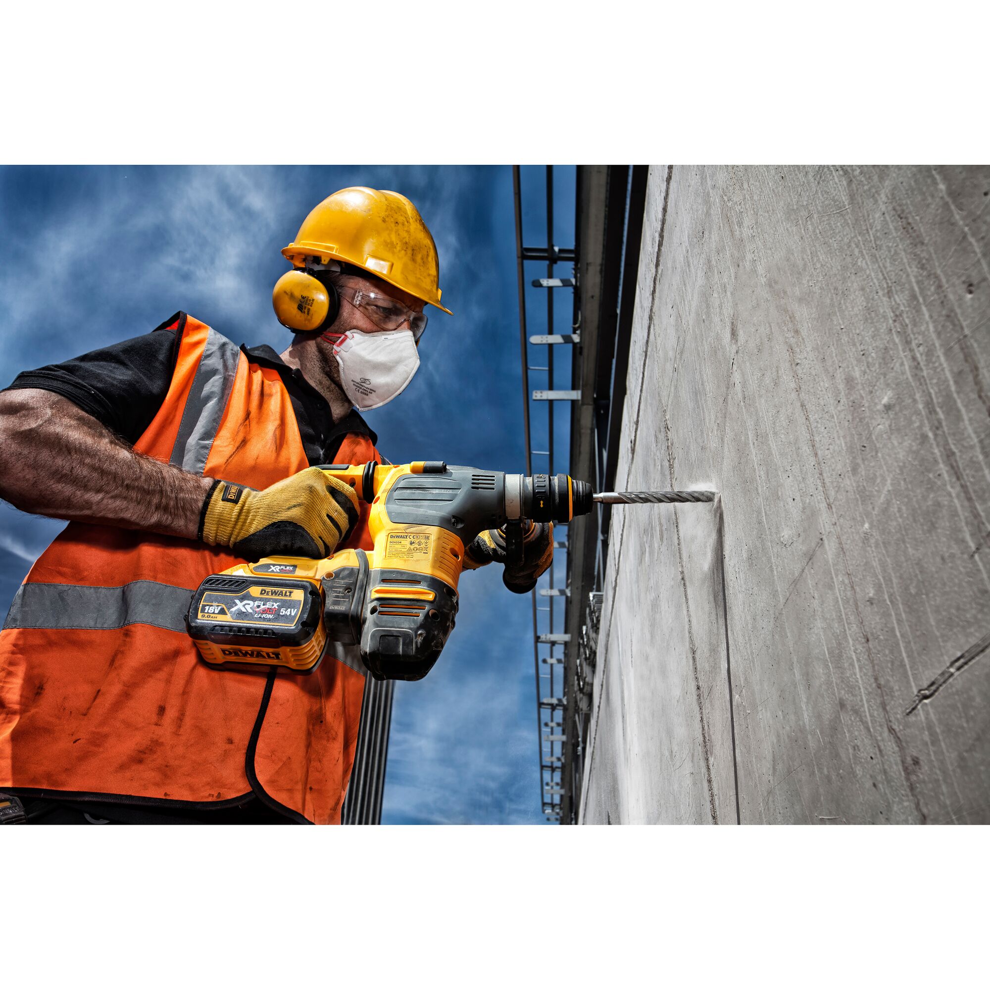 A construction worker wearing an orange safety vest, yellow helmet, and gloves is using a DEWALT cordless hammer drill to drill into a concrete wall outdoors under a blue sky.