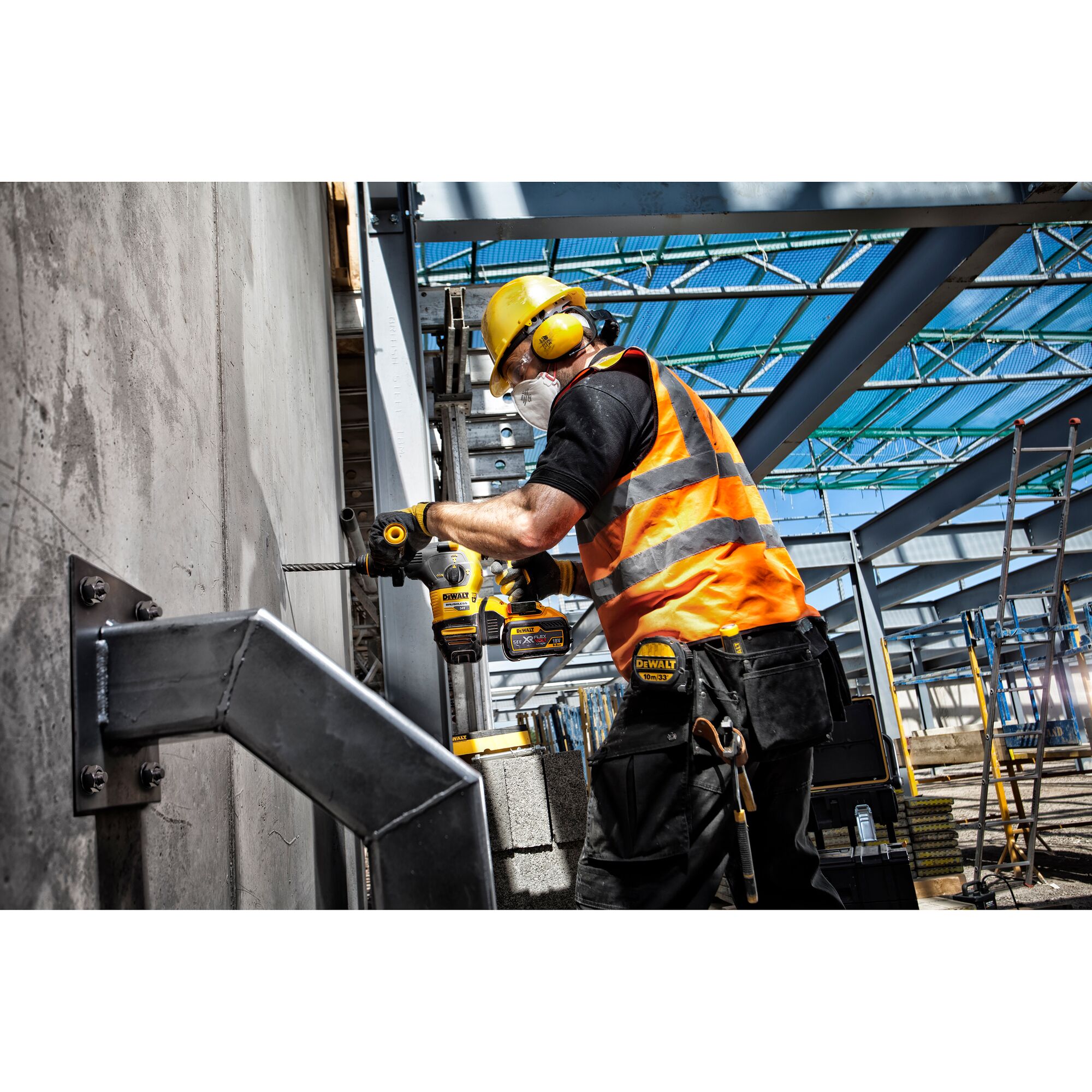 A construction worker wearing a yellow safety helmet, orange reflective vest, ear protection, and a dust mask is using a DEWALT power drill to drill into a concrete wall on a construction site.