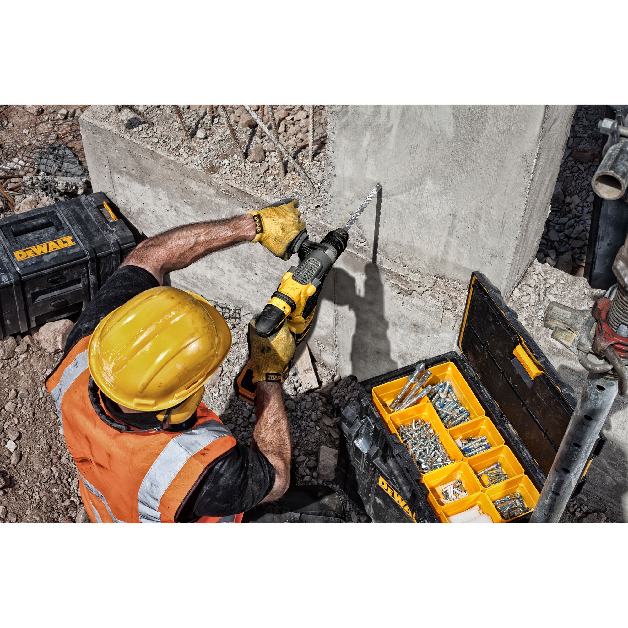 A construction worker wearing a yellow helmet and orange safety vest is using a DEWALT power drill to drill into a concrete wall. There are DEWALT toolboxes nearby, one of which is open and contains various hardware such as screws and anchors.