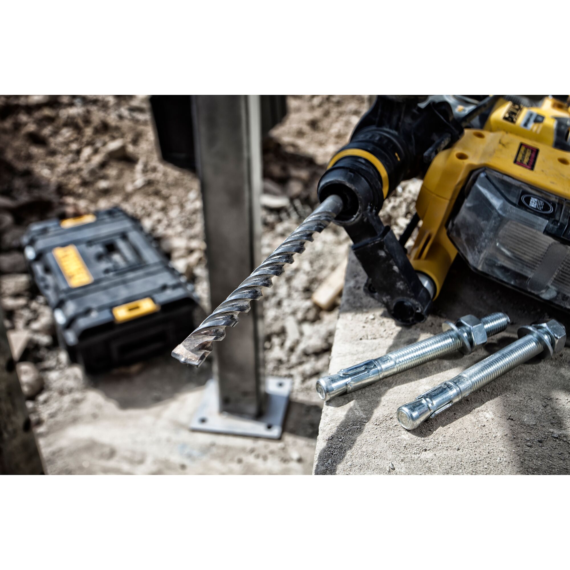 A close-up of a DEWALT rotary hammer drill with a spiral drill bit attached, resting on concrete. Three large metal anchor bolts are on the concrete surface nearby. In the background, a DEWALT tool case sits on rough ground at a construction site.