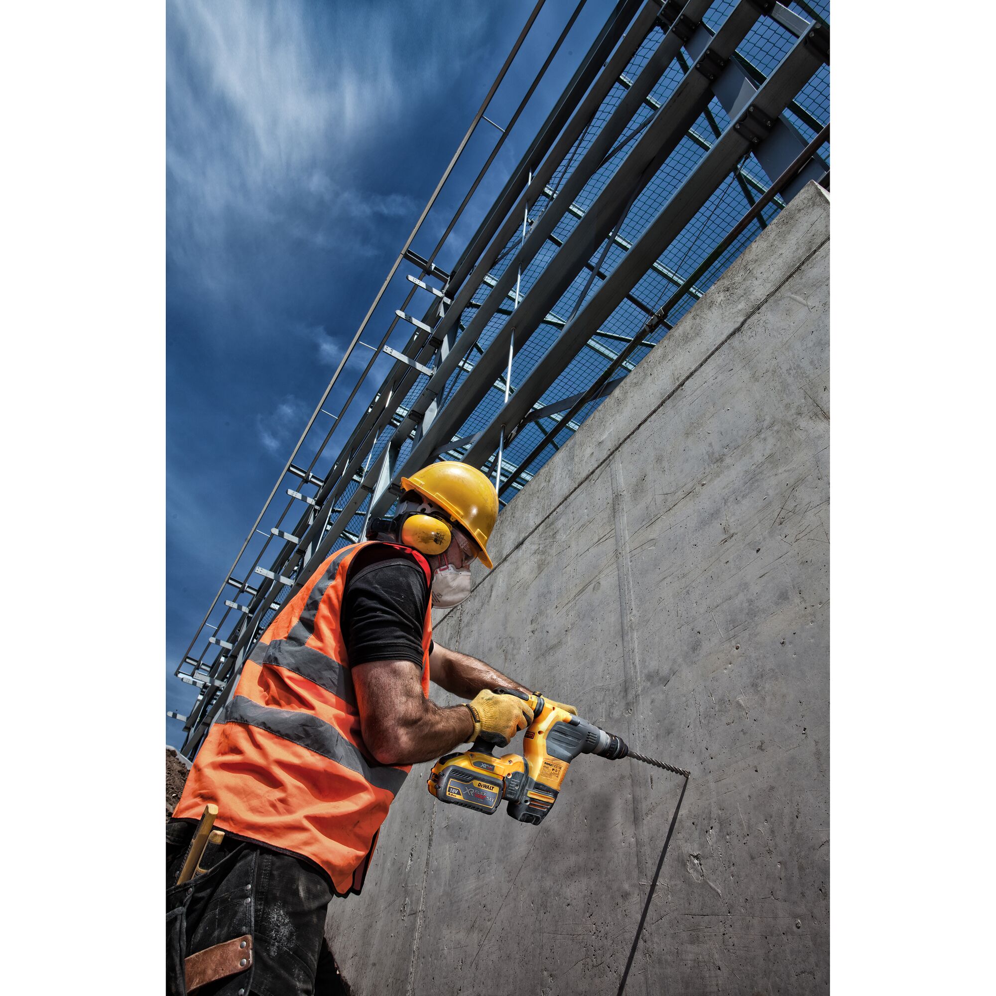 Construction worker wearing safety gear, including a yellow helmet, ear protection, safety vest, and gloves, using a DEWALT power drill to drill into a concrete wall outdoors on a sunny day.