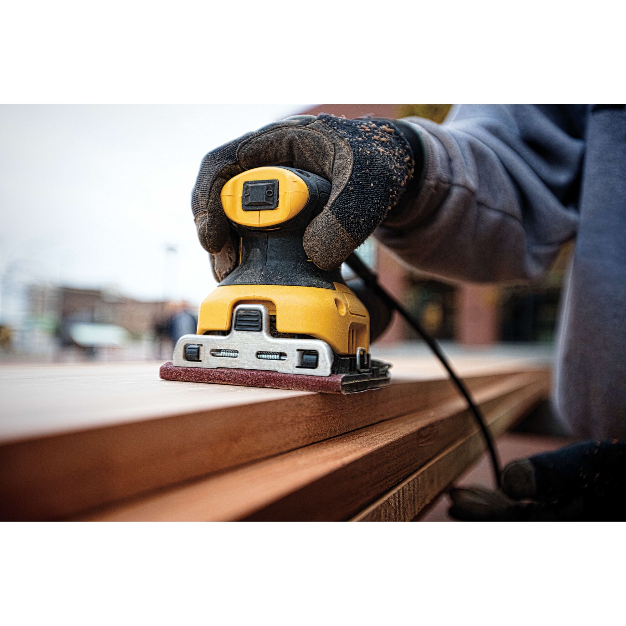 A close-up of a person using a DEWALT DWE6411 yellow and black electric sander to smooth a wooden surface outdoors. The person is wearing protective gloves.