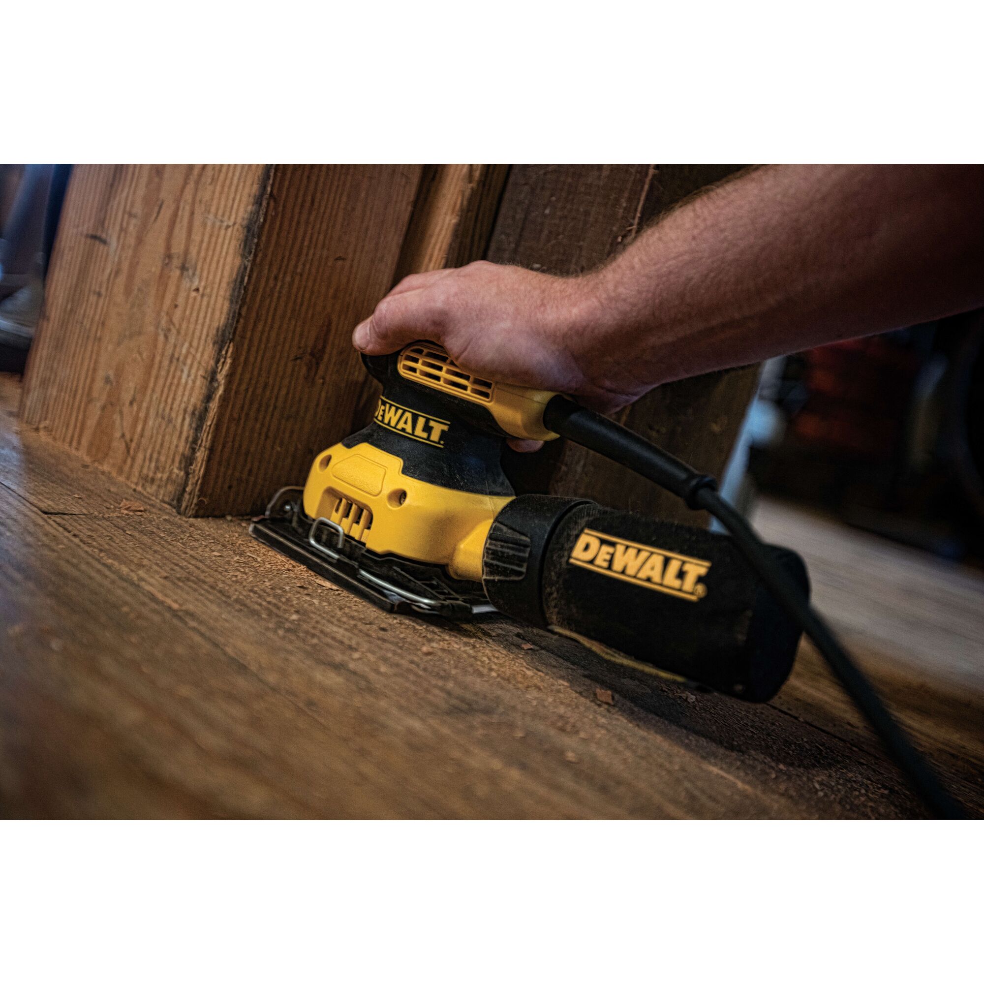 A close-up of a hand using a yellow and black DEWALT electric sander on a wooden floor next to a wooden post. The sander is plugged in and its DEWALT branding is clearly visible.