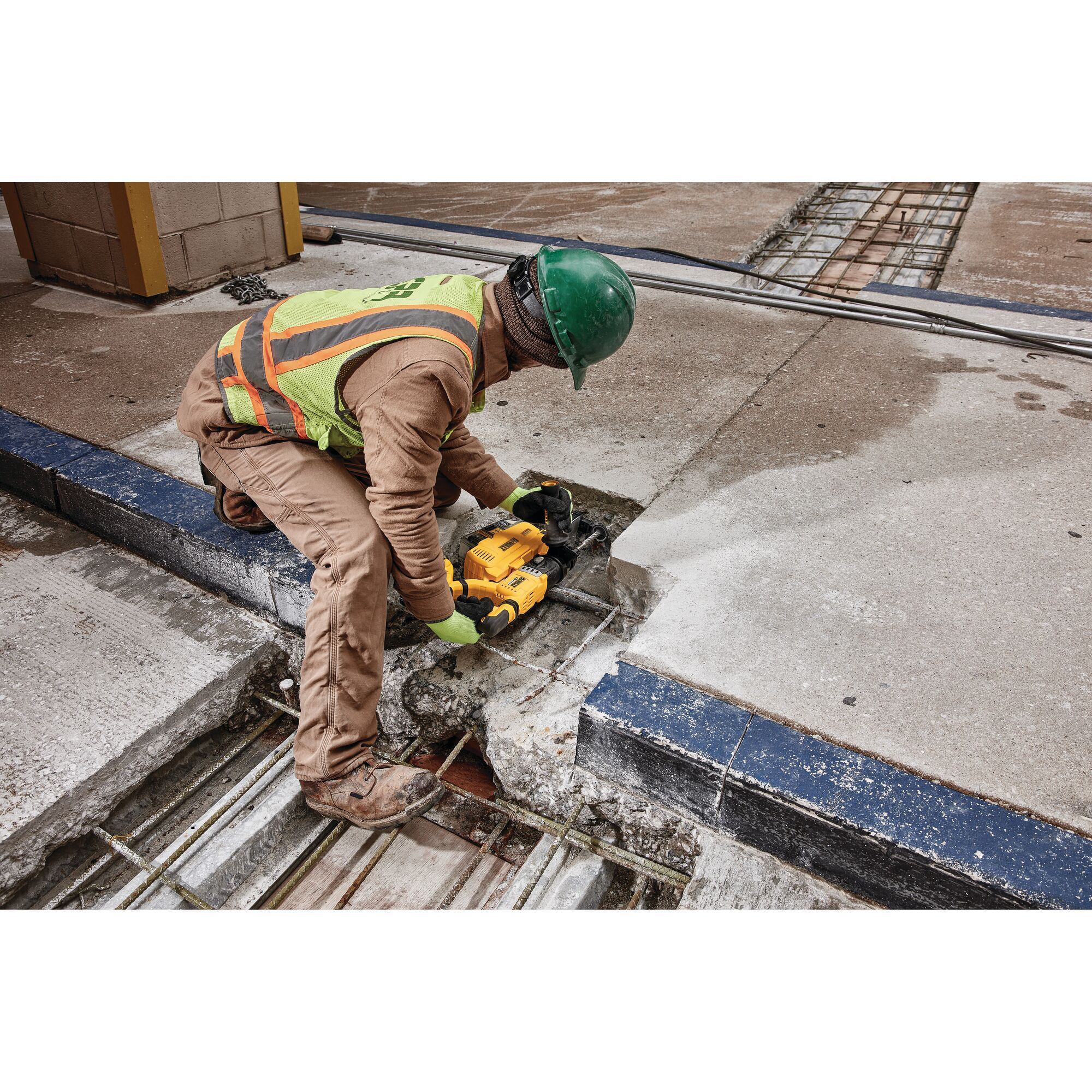A person wearing safety gear and a green helmet uses a DEWALT DWH205DH tool to cut into a concrete surface at a construction site. Rebar is visible underneath the concrete.